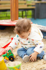 Fototapeta premium Baby girl boy in a white hoodie and jeans plays with a toy tractor. Happy Caucasian child 1 year old on a walk, vertical portrait