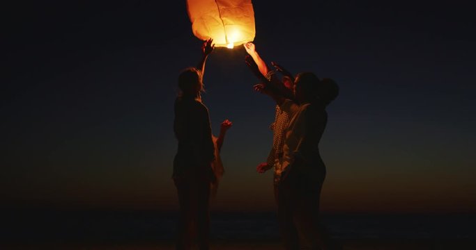 Slow motion silhouette of a group of young friends are having fun to letting go a sky lantern while standing on the beach on a sunset.