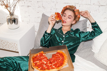 young fashionable woman in silk pajamas lays on bed and eat pizza pepperoni from pizza box, direct...