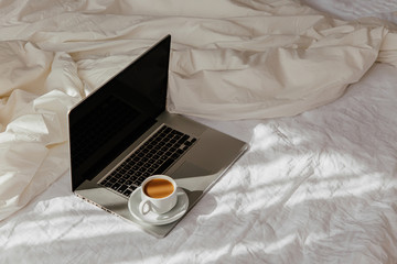 Fototapeta na wymiar Laptop and cup of coffee on white bed with a blanket. Work at home concept. Morning light