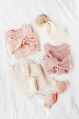 Obraz na płótnie Canvas Three pale pink and white warm sweaters and winter hat on bed. Women's stylish clothes. Cozy Winter look. Flat lay, top view.