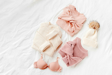 Fototapeta na wymiar Three pale pink and white warm sweaters and winter hat on bed. Women's stylish clothes. Cozy Winter look. Flat lay, top view.
