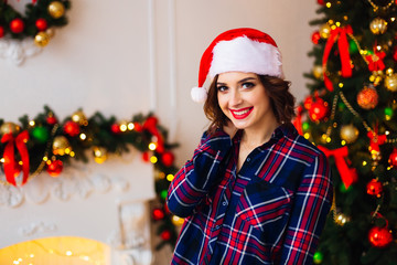 beautiful girl holds her hand in her hair with a santa clause hat on her head in a room with christmas tree and fireplace