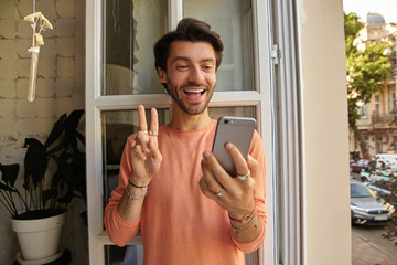Indoor shot of attractive young man with beard leaning on opened window while having videochat with his friends, smiling cheerfully to camera and showing peace gesture