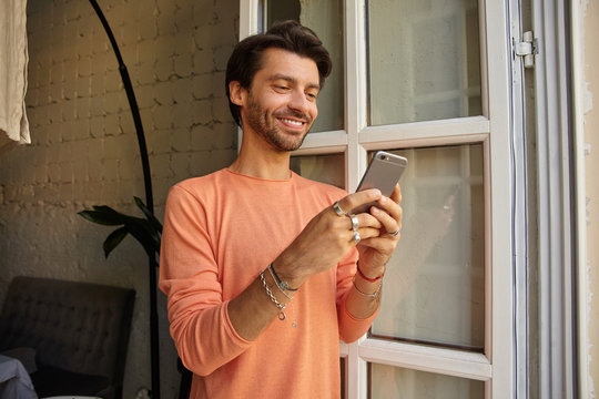 Indoor photo of positive young dark haired male in peach color sweater leaning on opened window, holding mobile phone in hands and looking at screen with wide smile