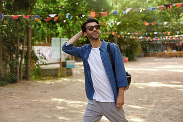 Obraz premium Beautiful young dark haired male in sunglasses walking through city park, looking upwards with broad smile and keeping hand on back of his head