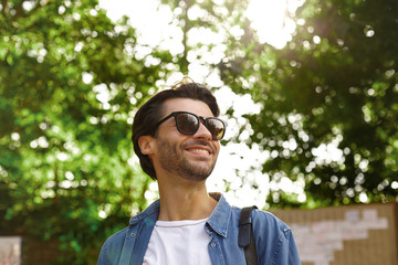 Outdoor portrait of happy pretty bearded male with dark hair looking aside and smiling cheerfully, posing over green trees on warm bright day