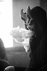 Vape man in devil mask and a hood smoking an electronic cigarette indoors. Puffs of steam. Bad habit that is harmful to health. Vaping activity. Black and white.