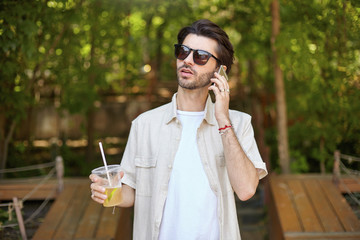 Young attractive bearded man in beige shirt and sunglasses making call with his mobile phone, walking around green city prk with lemonade in hand