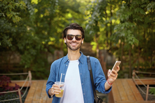Happy beautiful dark haired male in sunglasses looking at camera cheerfully and holding mobile phone in hand, wearing white t-shirt and blue shirt