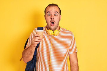 Student man wearing backpack headphones drinking coffee over isolated yellow background scared in...