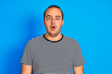 Young man wearing striped navy t-shirt standing over isolated blue background afraid and shocked with surprise expression, fear and excited face.