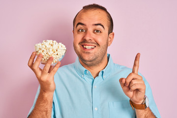 Young man holding bowl with popcorn standing over isolated pink background surprised with an idea or question pointing finger with happy face, number one