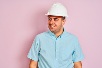 Young architect man wearing security helmet standing over isolated pink background looking away to side with smile on face, natural expression. Laughing confident.