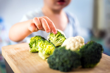 baby boy hands touch and take raw fresh broccoli and cauliflower from wooden board indoor. baby exploring vegetables