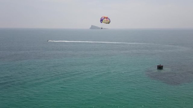 Colorful parasail wing pulled by sailing boat in turquoise Mediterran sea with big rock island in the background. Seagull flying across the camera on foreground.