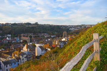 Fototapeta na wymiar Bird's eye view of a quintessential British coastal town with its charming traditional houses. Wooden fence on a lush grassy hill exposed to blue sky dotted with fluffy clouds. Hastings, England