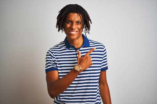 Afro man with dreadlocks wearing striped blue polo standing over isolated white background cheerful with a smile on face pointing with hand and finger up to the side with happy and natural expression