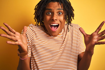 Afro man with dreadlocks wearing striped t-shirt standing over isolated yellow background very happy and excited, winner expression celebrating victory screaming with big smile and raised hands