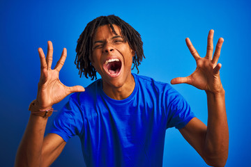 Afro american man with dreadlocks wearing t-shirt standing over isolated blue background celebrating mad and crazy for success with arms raised and closed eyes screaming excited. Winner concept