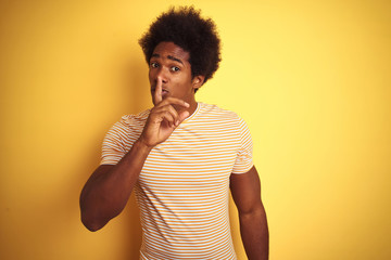 American man with afro hair wearing striped t-shirt standing over isolated yellow background asking to be quiet with finger on lips. Silence and secret concept.