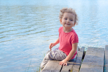 little cute girl sitting on a wooden pier on the shore, looking at the camera and smiling.