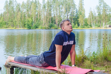 morning and evening, a handsome young man doing yoga exercise on the shore of a forest lake in nature in the summer outdoors. dog pose. healthy active lifestyle.