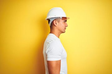 Young handsome man wearing construction helmet over yellow isolated background looking to side,...