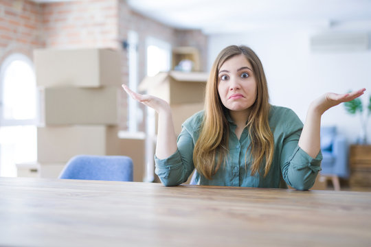 Young woman sitting on the table with cardboard boxes behind her moving to new home clueless and confused expression with arms and hands raised. Doubt concept.