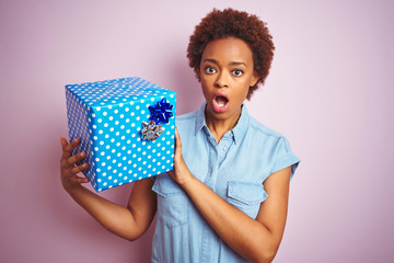 African american woman holding birthday gift over pink isolated background scared in shock with a...