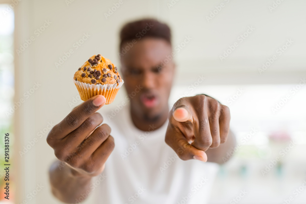 Poster African american man eating chocolate chips muffin pointing with finger to the camera and to you, hand sign, positive and confident gesture from the front