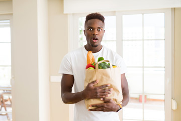 African american man holding paper bag full of fresh groceries scared in shock with a surprise face, afraid and excited with fear expression