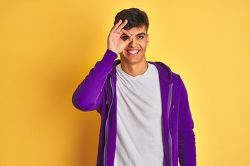 Young indian man wearing purple sweatshirt standing over isolated yellow background doing ok gesture with hand smiling, eye looking through fingers with happy face.