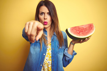 Young beautiful woman eating fresh healthy watermelon slice over yellow background pointing with...
