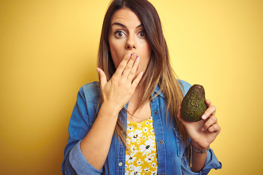 Young beautiful woman eating healthy avocado over yellow background cover mouth with hand shocked with shame for mistake, expression of fear, scared in silence, secret concept