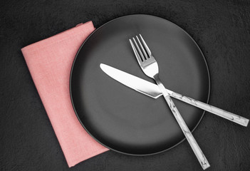 Black plate, cutlery and napkin on stone table top view. Table setting.