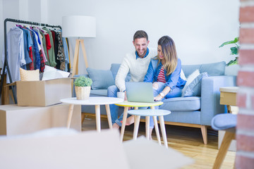 Young beautiful couple sitting on the sofa drinking cup of coffee using laptop at new home around cardboard boxes