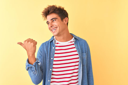 Young handsome man wearing striped t-shirt and denim shirt over isolated yellow background smiling with happy face looking and pointing to the side with thumb up.
