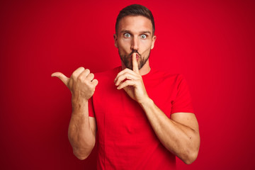 Young handsome man wearing casual t-shirt over red isolated background asking to be quiet with finger on lips pointing with hand to the side. Silence and secret concept.