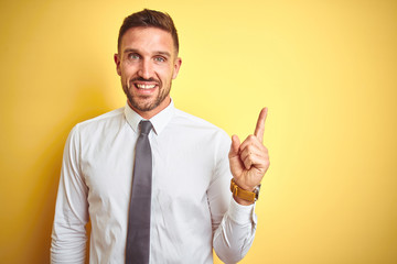 Young handsome business man wearing elegant white shirt over yellow isolated background with a big smile on face, pointing with hand and finger to the side looking at the camera.
