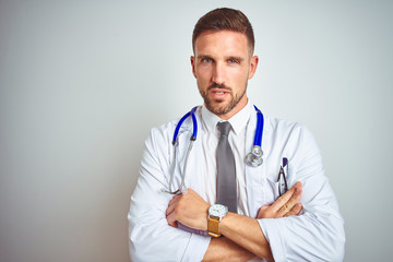 Young handsome doctor man wearing white professional coat over isolated background skeptic and...