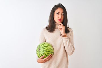 Young beautiful chinese woman holding watermelon standing over isolated white background serious...