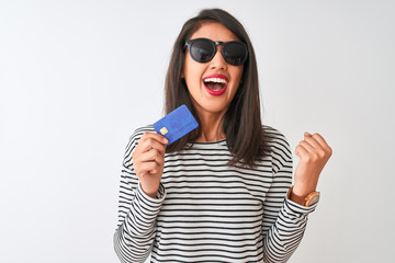 Young chinese woman wearing sunglasses holding credit card over isolated white background screaming proud and celebrating victory and success very excited, cheering emotion