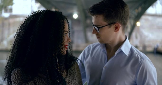 A close-up portrait of a young beautiful couple-a burning curly brunette girl and a young man in a shirt and glasses. They stand under the bridge, romantic look at each other, smiling, hugging.
