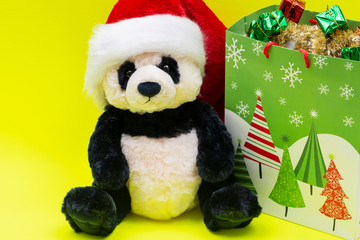 Bag with Gifts and Panda Bear plush toy wearing Red Christmas Santa Hat on bright yellow background. Christmas, New Year, Winter Holidays concept.