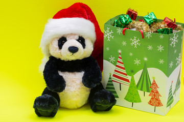 Bag with Gifts and Panda Bear plush toy wearing Red Christmas Santa Hat on bright yellow background. Christmas, New Year, Winter Holidays concept.