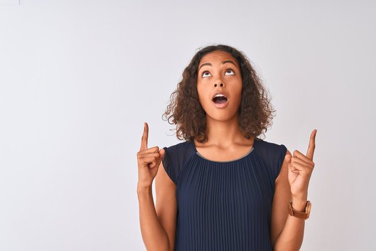 Young brazilian woman wearing blue dress standing over isolated white background amazed and surprised looking up and pointing with fingers and raised arms.