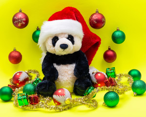 Panda Bear plush toy with Red Christmas Santa Hat surrounded by Tinsel Garland and Vintage Ornaments on bright yellow background. Holidays concept.