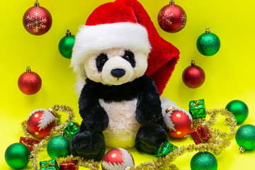 Panda Bear plush toy with Red Christmas Santa Hat surrounded by Tinsel Garland and Vintage Ornaments on bright yellow background. Holidays concept.