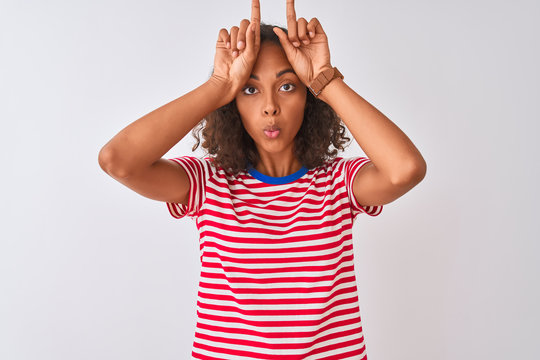 Young brazilian woman wearing red striped t-shirt standing over isolated white background doing funny gesture with finger over head as bull horns